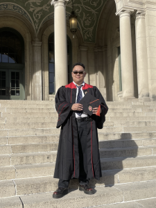 A man wearing sunglasses and a black graduation robe, smiles.